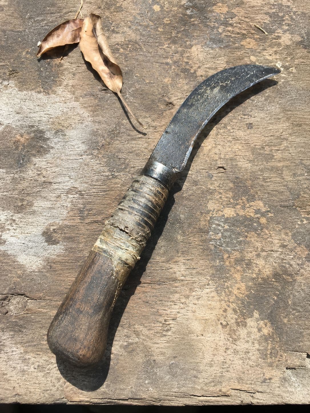 A curved grafting knife with a worn wooden handle resting on a stone surface with two dry leaves.