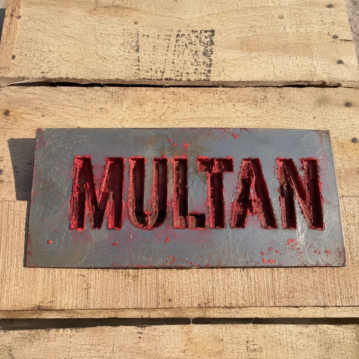 A metal stencil plate cut with the word MULTAN, resting on a timber board and inked red.