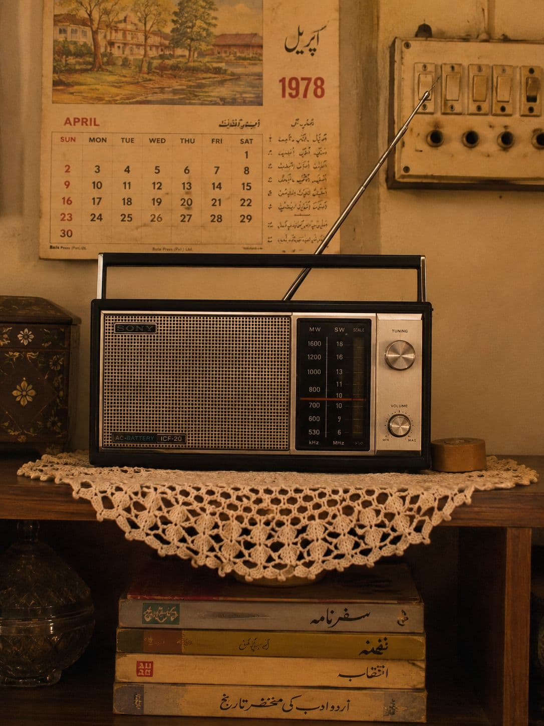 A transistor radio on a crocheted doily above a stack of Urdu paperbacks, beside a wall calendar.